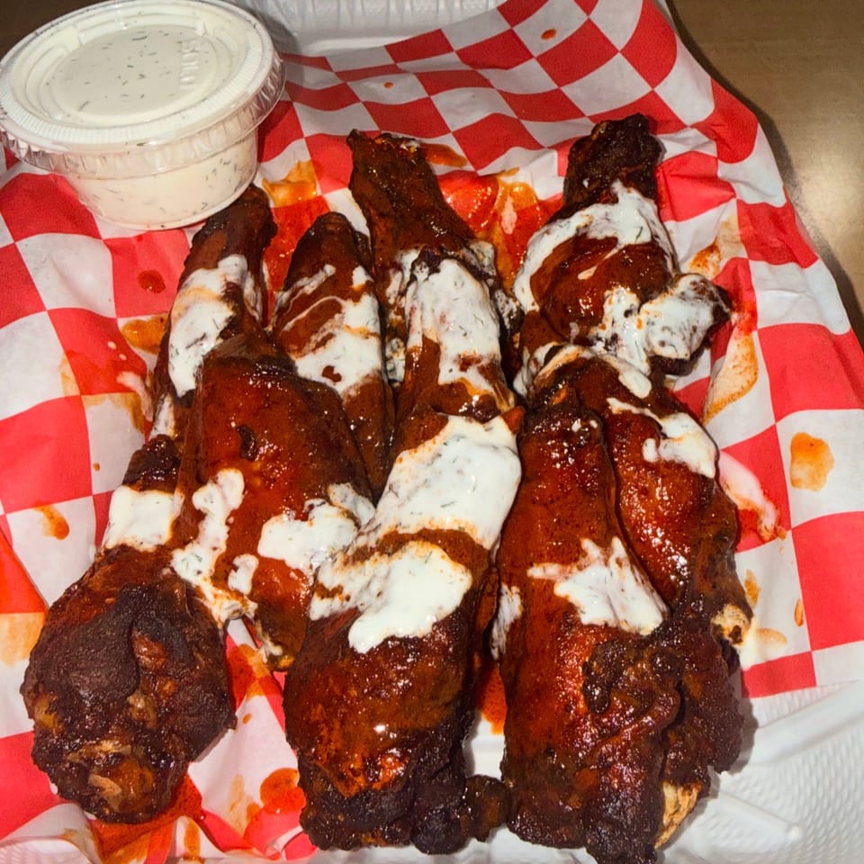 Glazed grilled chicken wings drizzled with white sauce in a red-and-white checkered takeout basket, with dipping sauce.