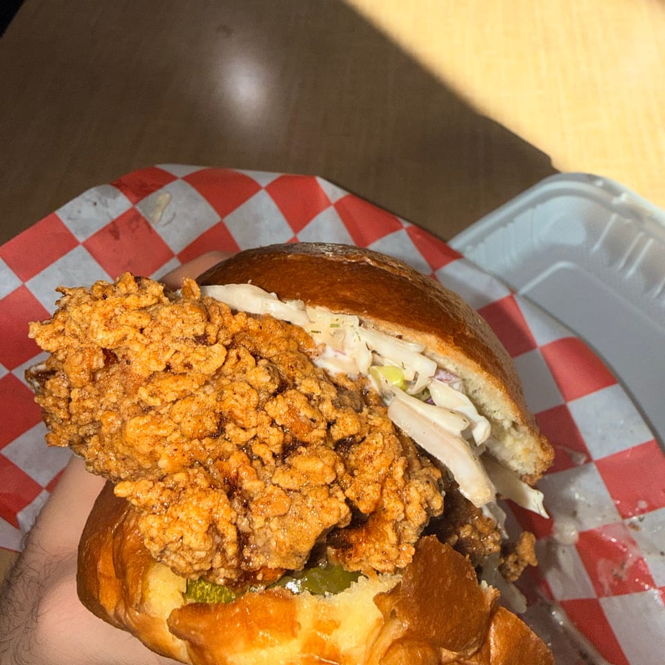 Close-up of a crispy fried chicken sandwich with coleslaw, pickles, and sauce on a toasted bun in a checkered basket liner.