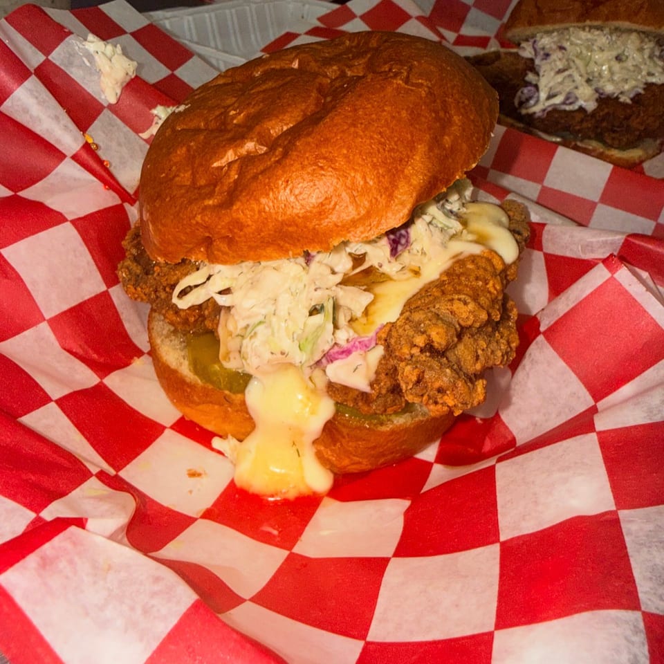 Crispy fried chicken sandwich with coleslaw, pickles, and melted cheese on a brioche bun, served on red-and-white checkered paper.